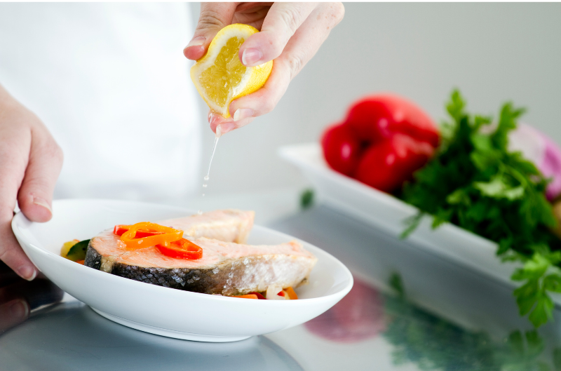 person squeezing fresh lemon on fish with vegetables in the background